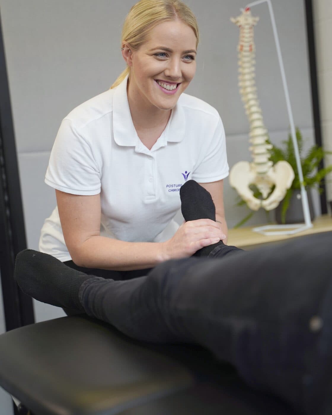 Bianca Downey laughing as she holds a patient's ankle on the treatment table, the spine model behind her.