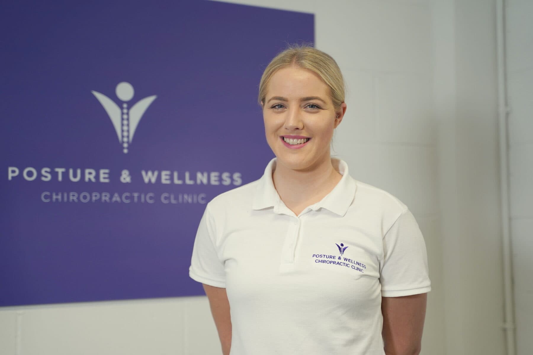 Bianca Downey smiling in front of the purple Posture & Wellness sign at the clinic.
