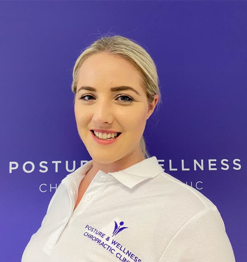 Bianca Downey in her Posture & Wellness polo, against the purple clinic sign.
