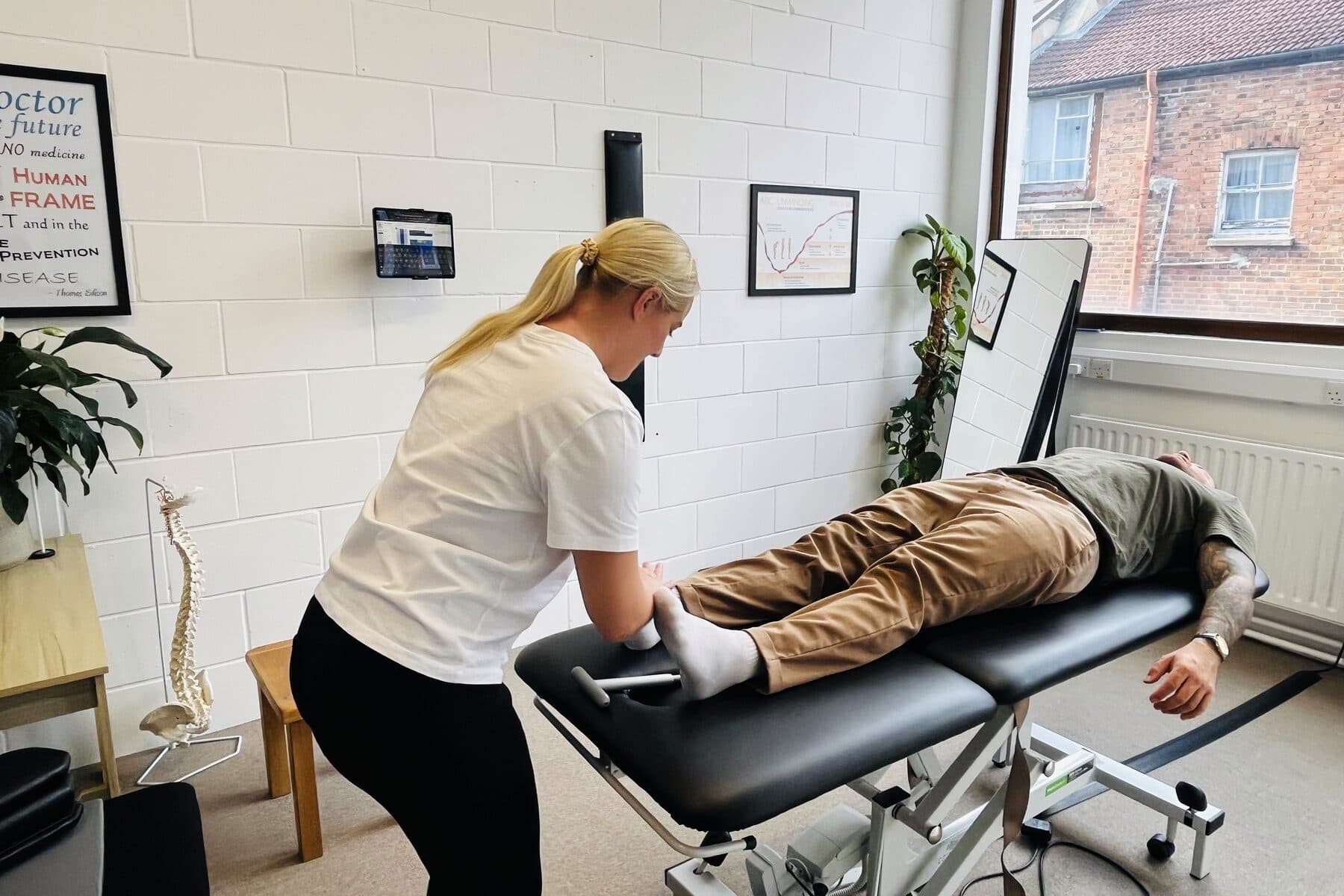 The treatment room: Bianca Downey working at a patient's feet on the table, the spine model and wall charts behind.